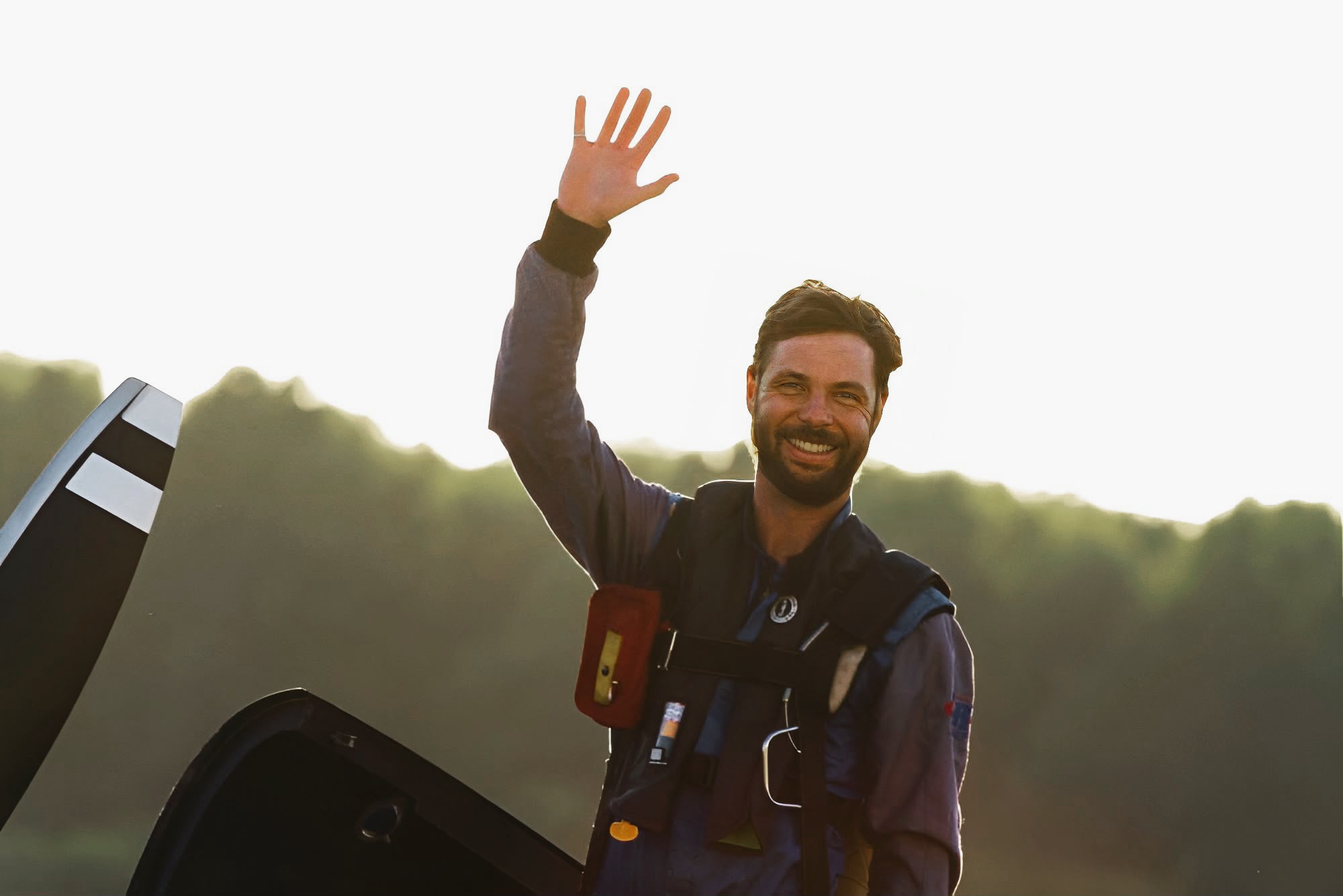 Mike Tryggvason in a flight suit and life vest, waving from the open cockpit of his aircraft, backlit by low sun over a treeline